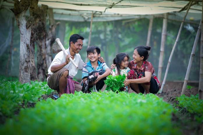 A family kneels amidst rows of bright green vegetables. Their arms are around one another. They are smiling broadly.