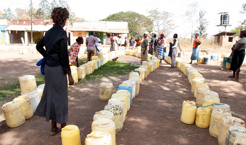 Jerry cans are lined up on the ground in a village and people stand around them.