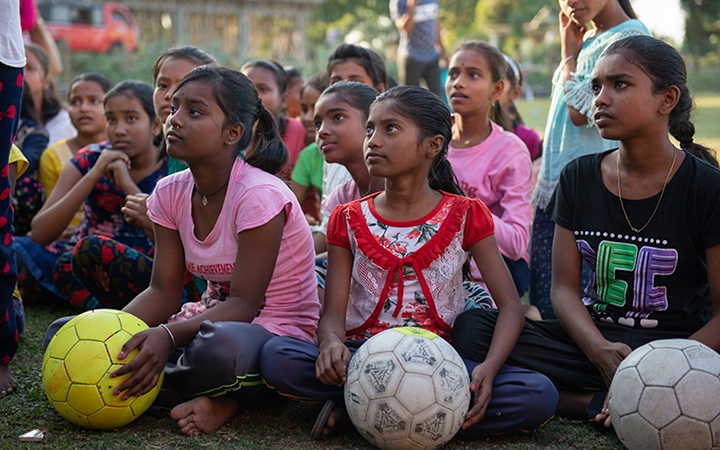 A group of adolescent Indian girls sits cross-legged on grass looking up. The three girls in the front row hold soccer balls.