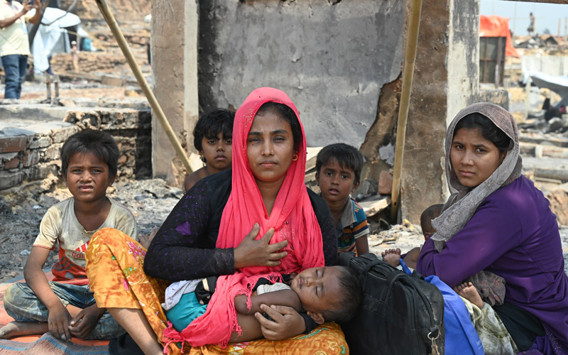 two Rohingya women sits on the ground surrounded by small children in the midst of a burned out temporary structure.