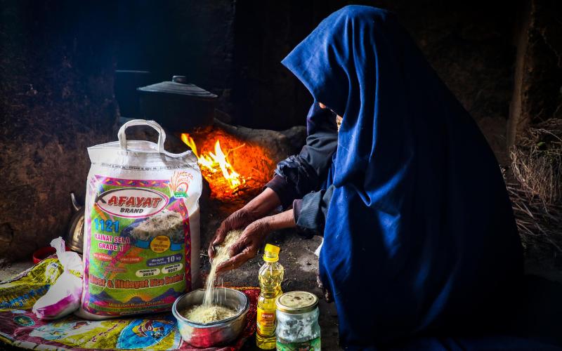 An Afghan woman wearing a rich, blue hijab squats to prepare rice on a concrete floor.