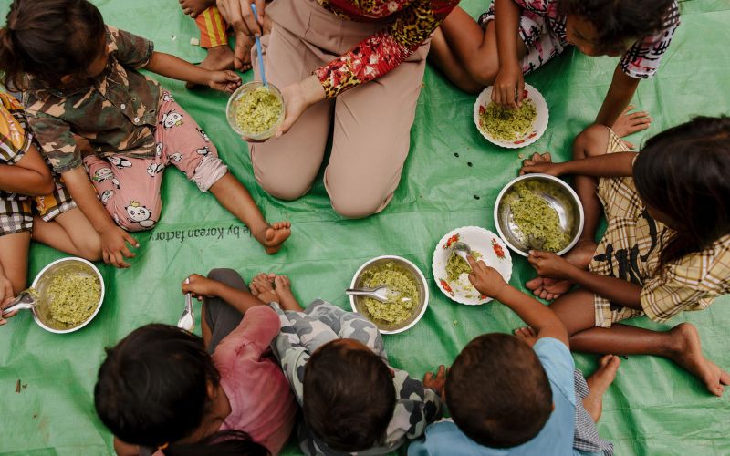 A group families eat food from bowls together, sitting on a mat.