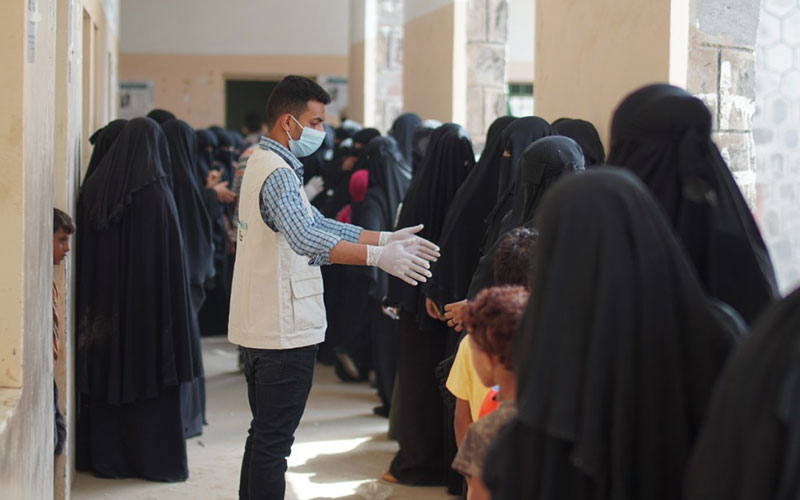 An aid worker wearing a mask indicates the need for physical distancing in a long lineup of women wearing black head coverings and garments.