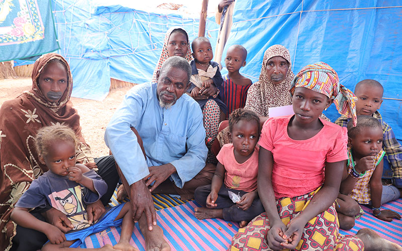 A large family group in Mali sits together on the ground, in a refugee camp setting.