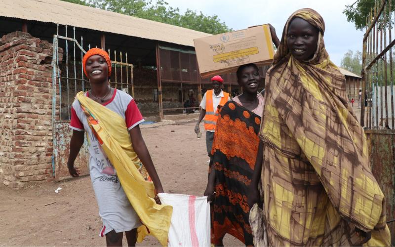 Three smiling women exit a compound in Malakal, South Sudan, carrying bags and boxes of food.