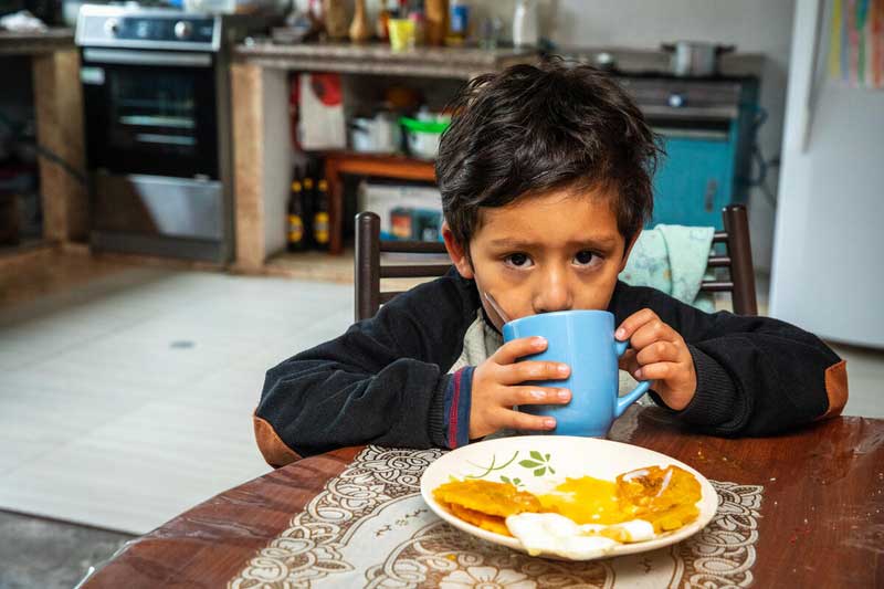 A boy wearing an elbow patch sweater sits at a dining table, drinking from a blue cup as part of his morning breakfast in Ecuador.