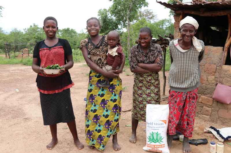 Four women and a child stand smiling next to their home in Zambia.