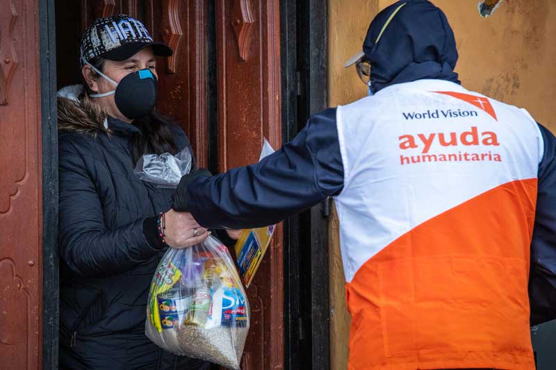 A woman in a black baseball cap and protective mask receives a care kit from a World Vision Ecuador staff member at her front door.