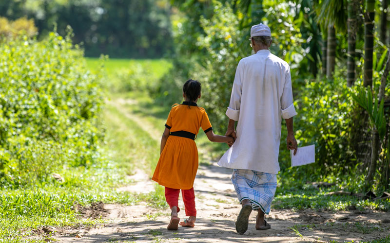 a young Bangladeshi girls walks down a green path with her grandfather.