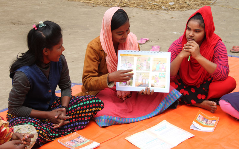 three Bangladeshi girls sit on the ground. they are looking at a booklet.