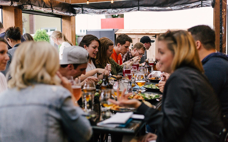 A group of people are seated, eating and chatting around a table filled with food and drink on an outdoor patio.