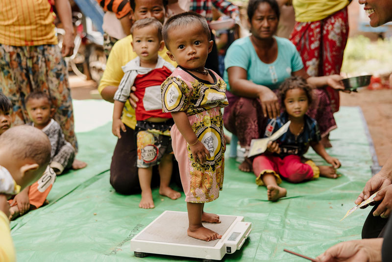 An infant child stands on a scale to have their weight taken. A crowd of mothers and their children sit around awaiting their turn.