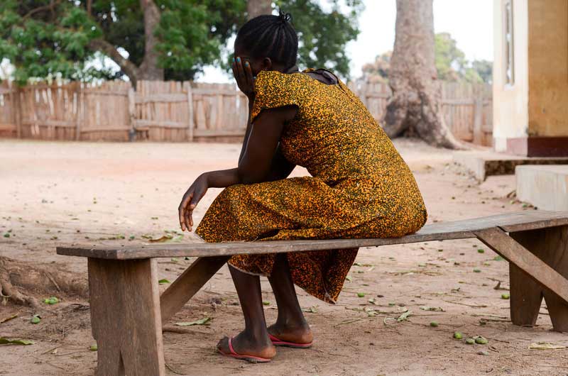 A woman from South Sudan wearing a yellow dress sitting on a wooden bench with her back to the camera.