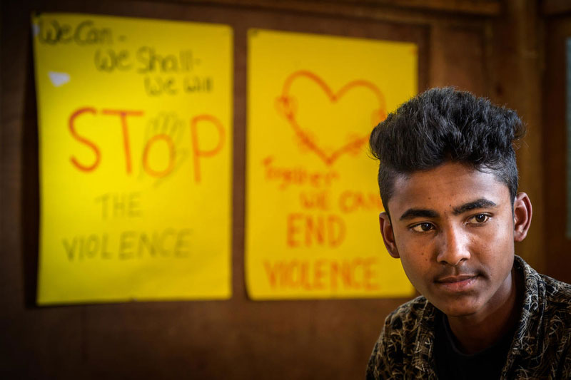 A Rohingya teenage boy sitting in front of a wall that has two large posters attached to.