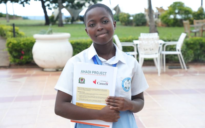 A young Tanzanian girl holds a booklet with the title "AHADI Project" over top, smiling at the camera.