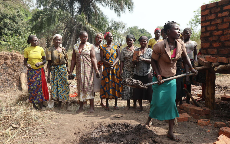 Mikelina shovels a pile of dirt, as her farmer's group of women look on, smiling.