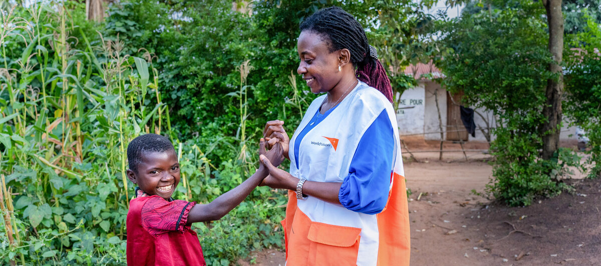 A smiling woman in a World Vision uniform playfully holds hands with a smiling young boy while standing in a clearing surrounded by crops.