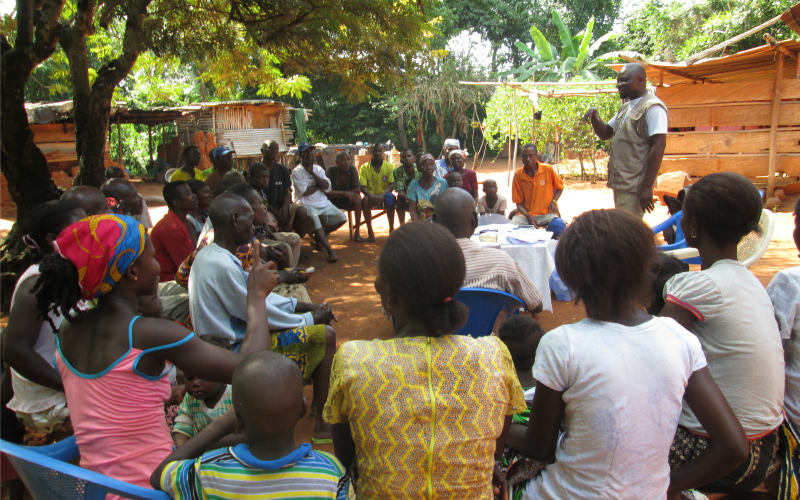 A man speaks to a community group outside under some trees