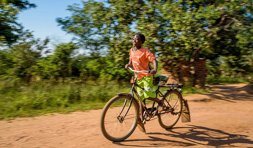 A child in orange shirt and green shorts speeds past trees and bushes on a bicycle on a dirt road