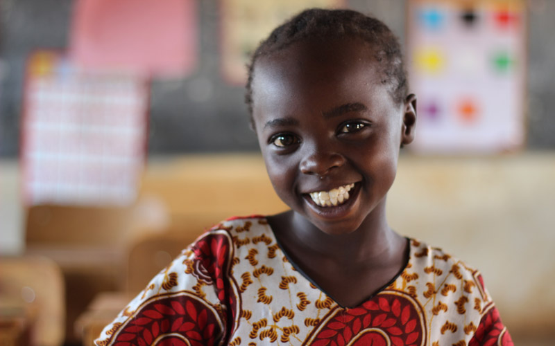 A young Kenyan girl sits in a classroom and smiles at the camera