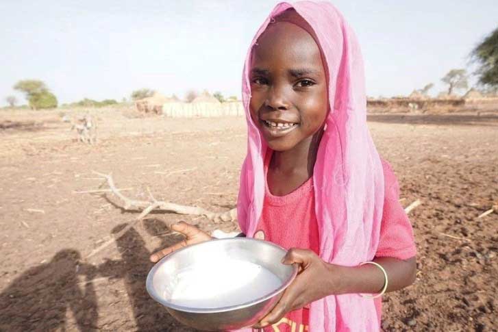 A small girl in Sudan wearing a pink headscarf stands on a parched landscape, holding a bowl of fresh, white goats’ milk. She is smiling.