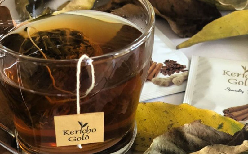 A cup of tea with a Kericho Gold tag. Dried tea leaves are scattered around it.