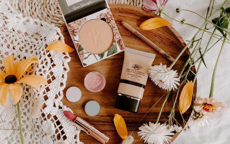 Makeup products laid out on a round wooden plate, surrounded by flowers and a white cloth.