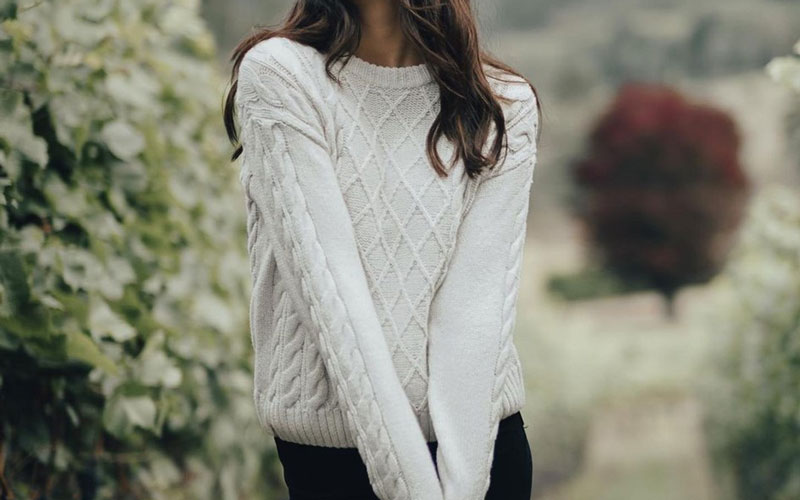 A woman in a farm, wearing a white wool cable crewneck sweater and a pair of black pants.