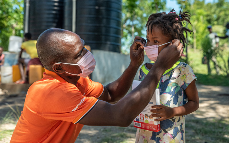 A Haitian man helps a small child on with a mask.