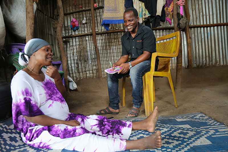 A man sits on a chair talking to a pregnant woman, wearing a white and purple dress. She sits on the floor, on a mat, in her home in a refugee camp in Kenya.