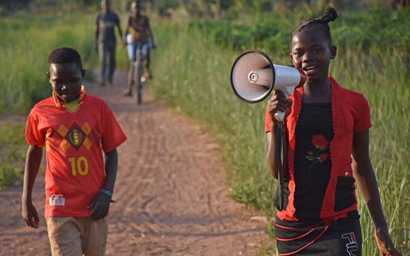 A young girl walks down a path holding a bullhorn up to her mouth.