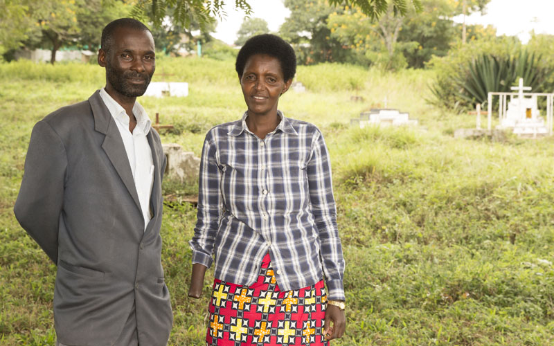 A man and a woman from Rwanda stand together under a tree in a graveyard.