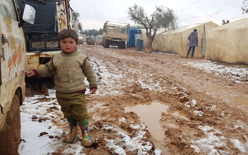 a young Syrian boy stands in the mud and snow at an IDP camp.
