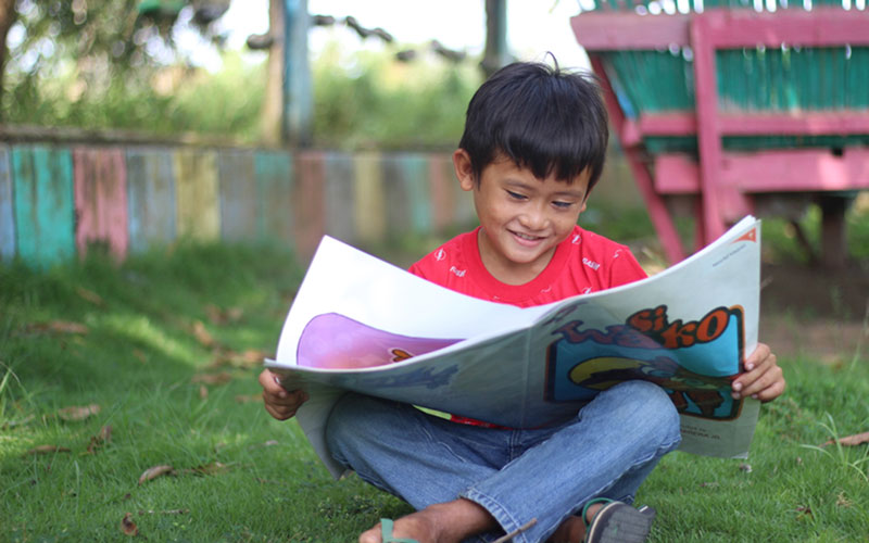 A little boy in the Philippines sits on the grass and reads a large publication for children.