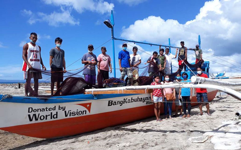 A group of adults and children stand on and near a fishing boat on a beach in the Philippines.