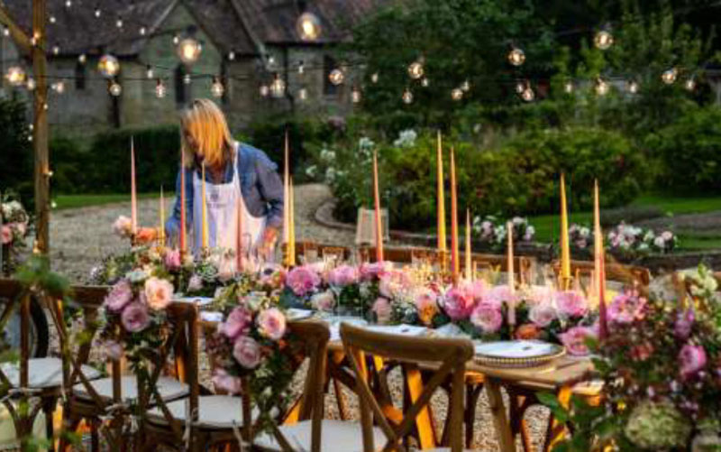 A woman prepares a long table for a meal, outdoors. The table is decorated with pink roses.