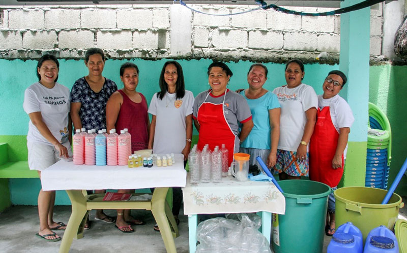 A group of smiling women stands behind tables with bottles of water and of soap.