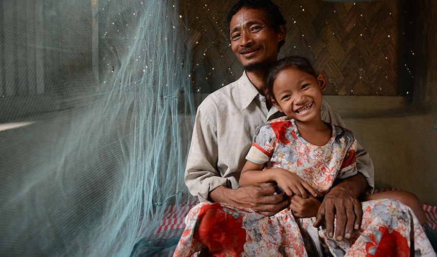 Father holds smiling daughter in his lap with mosquito net in the background