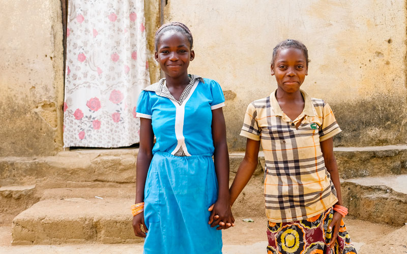 two girls from Central African Republic smile outside their home, and hold hands.