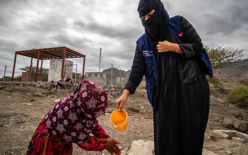 In Yemen, a little girl bends to wash her hands from a small jug of water poured by a woman. Around them, the landscape is bleak.
