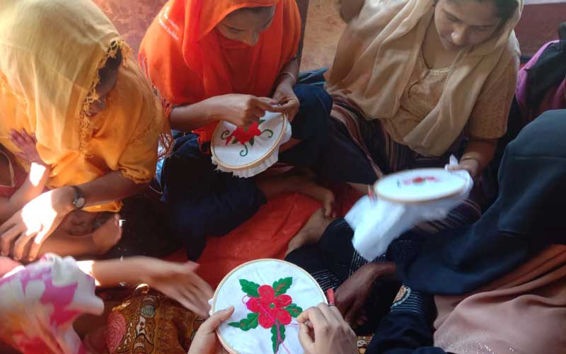 A group of women and older girls wearing headscarves sit in a circle, doing needlework together.