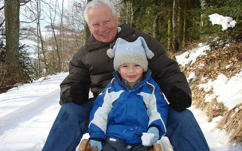 A grandfather and grandson prepare to go sledding down a hill in the winter.