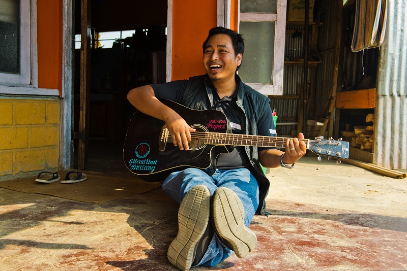 A man laughs while sitting on the floor playing guitar.