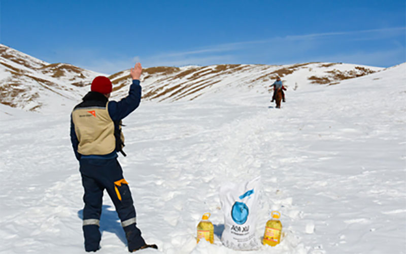 A World Vision worker in snow pants and a wool hat waves to a man riding an animal out of the mountains.