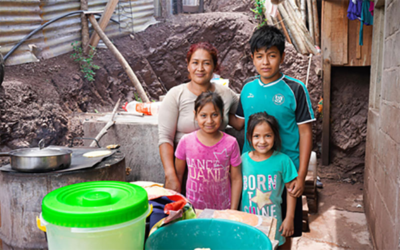A mother and her three school-age children stand in front of some buckets, outside their corrugated tin home.