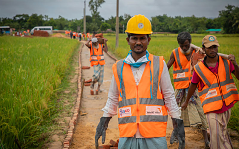 A group of men wearing reflective orange vests and hard hats work to repair a road in Bangladesh.