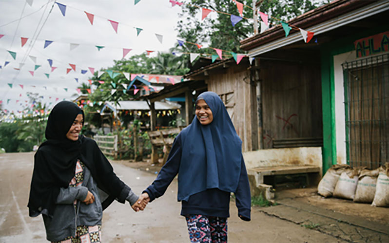 Two adolescent girls walk through the streets of their community, holding hands and laughing.