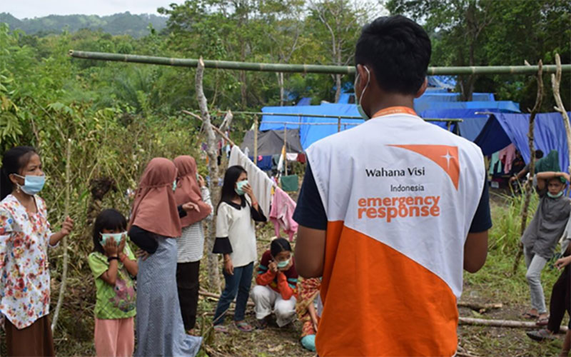 A World Vision worker with a mask on talks to families who are building temporary shelters, with bamboo poles and tarpaulins.