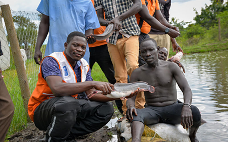A group of men in Uganda stand and sit beside a pond, with a World Vision worker.
