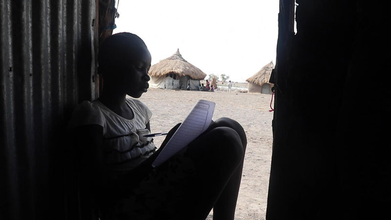Nyangieth sits in the doorway of her home, writing on a worksheet, with the background of her neighbours’ huts behind her.